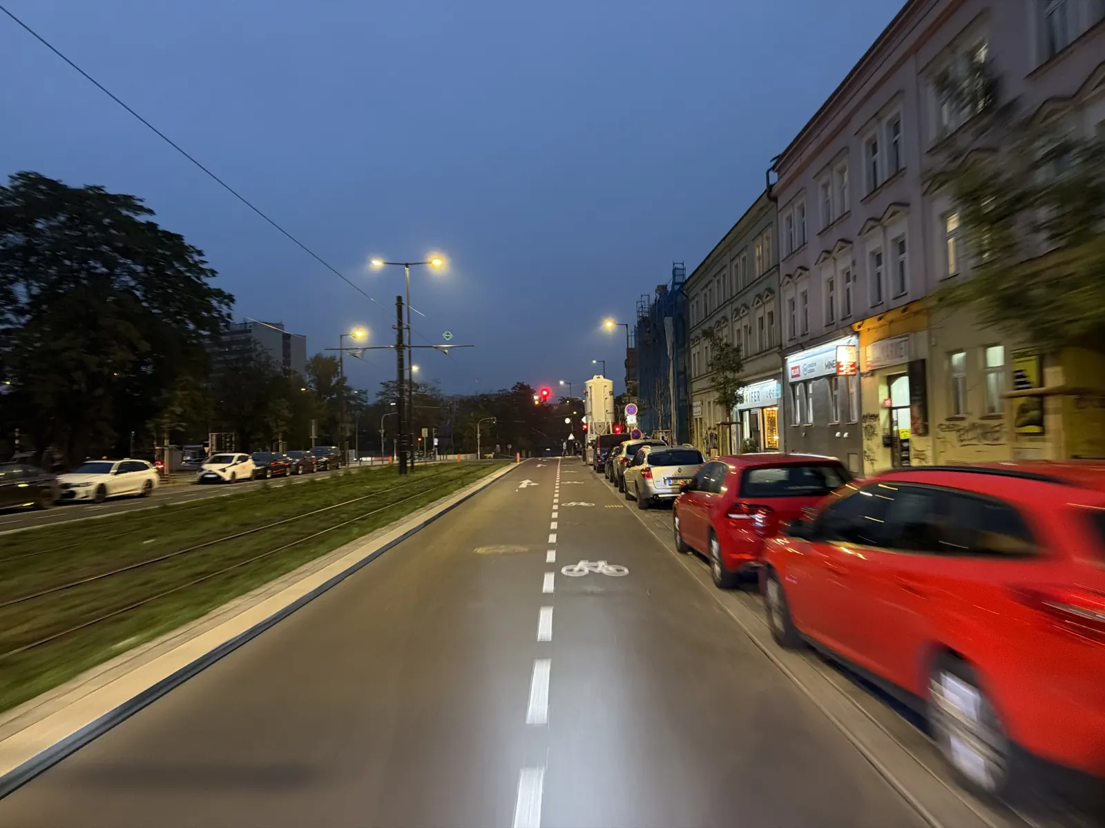 Vyznačený cyklistický pruh podél zaparkovaných aut, vlevo zatravněné tramvajové těleso.