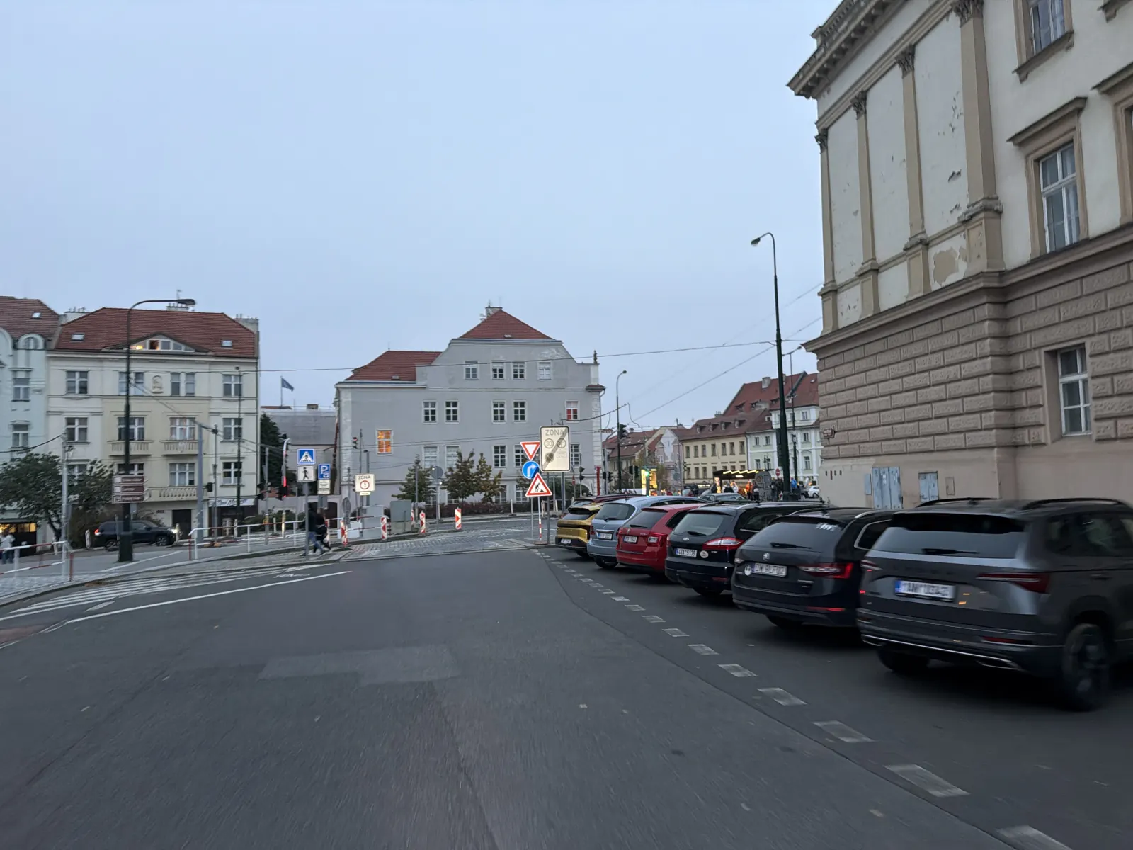 Městská ulice s řadou zaparkovaných aut, křižovatkou a dopravními zábranami