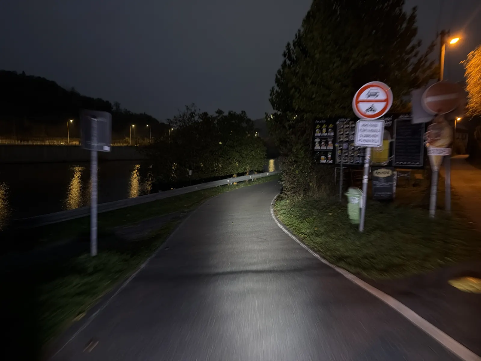 Cesta se stáčí podél řeky kolem občerstvení; na hladině se odrážejí světla.