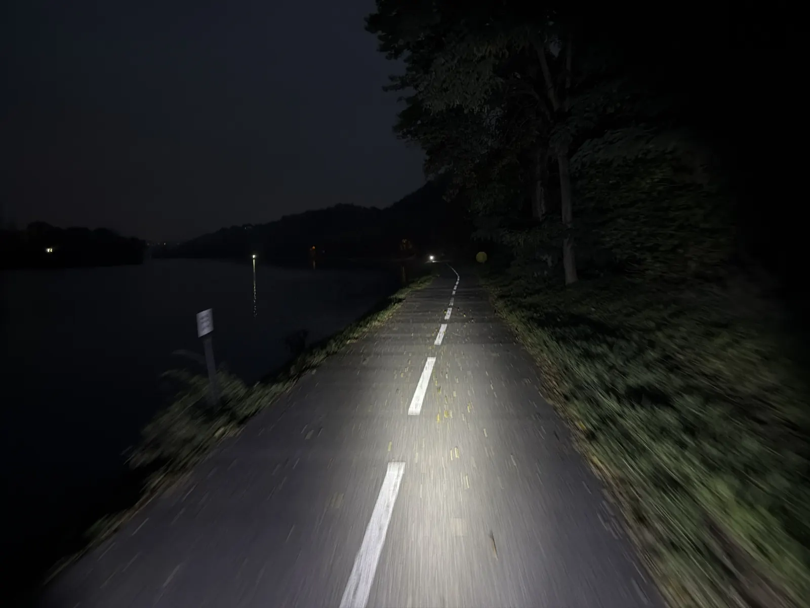 Noční stezka podél řeky s přerušovanou středovou čárou, osvětlená reflektorem.