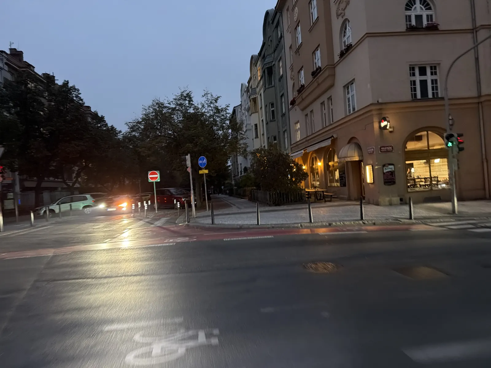 Večerní městská křižovatka s osvětlenou restaurací, cyklistickým značením a odlesky světel na vozovce